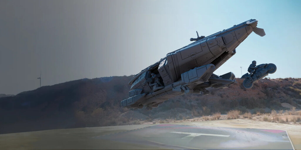 An unpainted model of a Herakles Aerodyne Vehicle hovering over a landing pad while a model of a bike and rider (again, unpainted grey) jumps out of the rear exit, ready to land below. A door gunner model can be seen covering the area. The scenery in the background is photo-realistic, in contrast to the models.